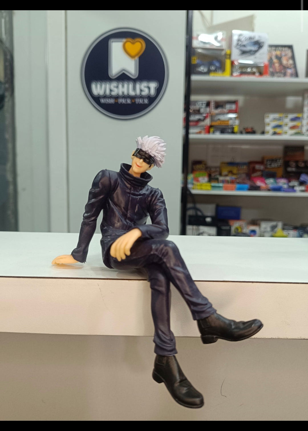 A collectible anime figurine of Satoru Gojo from Jujutsu Kaisen sitting on a white counter in a store, with a 'Wishlist' sign and shelves in the background.